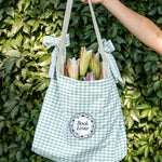 Green gingham tote bag with 'Book Lover' label held against a green leafy background