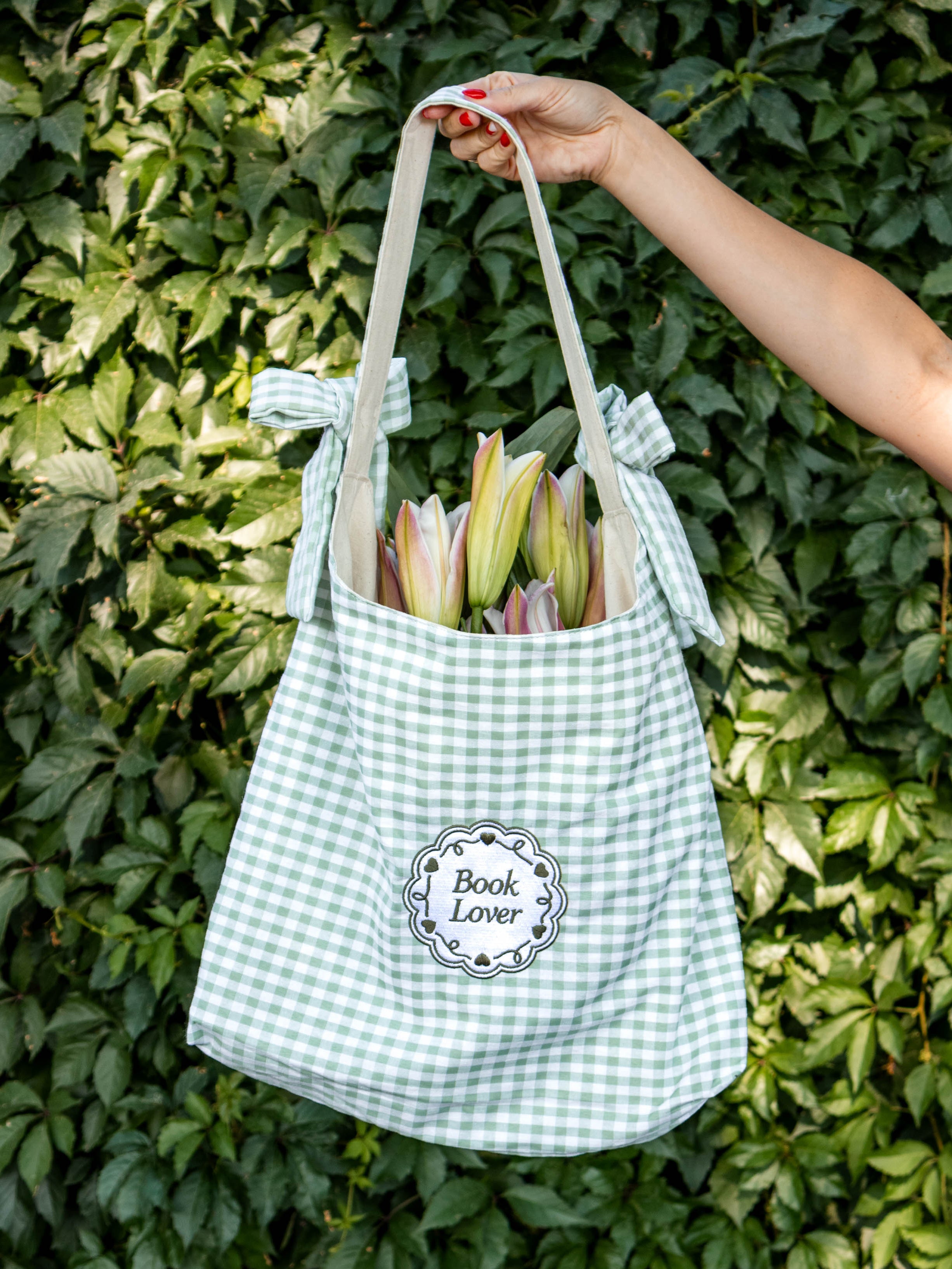 Green gingham tote bag with 'Book Lover' label held against a green leafy background