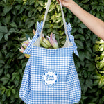 Blue and white checkered tote bag with 'Book Lover' text held against a green leafy background