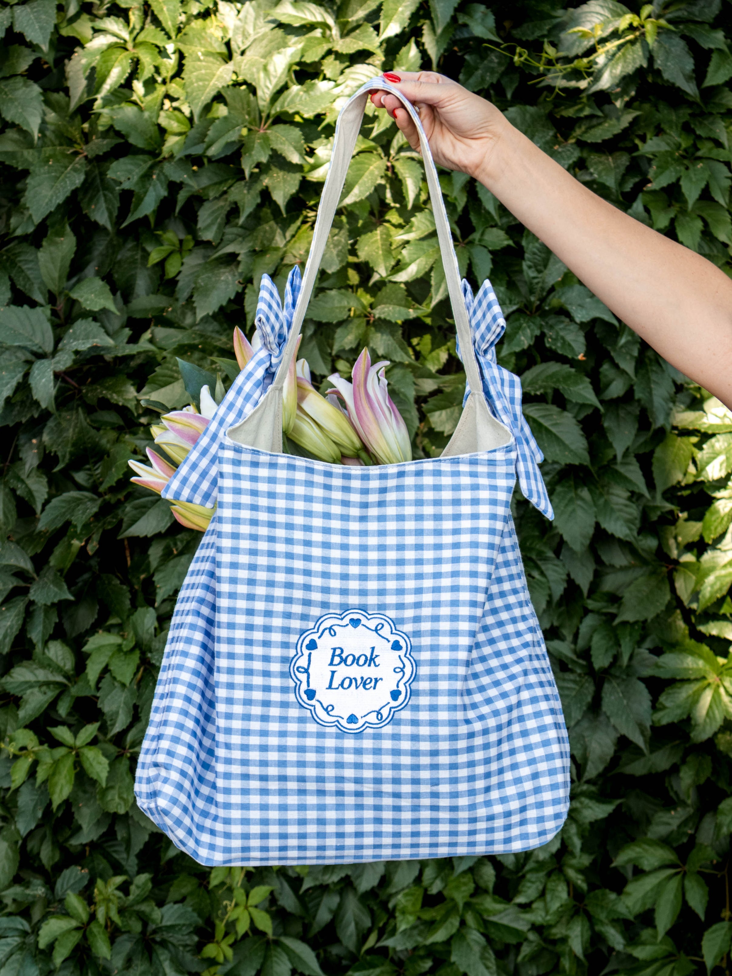 Blue and white checkered tote bag with 'Book Lover' text held against a green leafy background